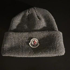 Grå stickad mössa från Moncler - Snygg grå stickad mössa från Moncler med broderad logotyp framtill. Perfekt för kyliga dagar med sin ribbade design som ger en skön passform.