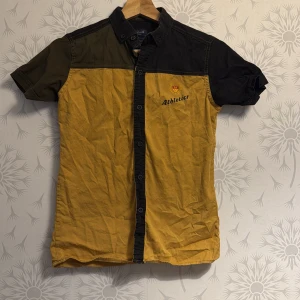 Gul och svart kortärmad skjorta - Snygg kortärmad skjorta i gult och svart från Athletic. Skjortan har knappar framtill och ett tryck på bröstet. Perfekt för en avslappnad stil.