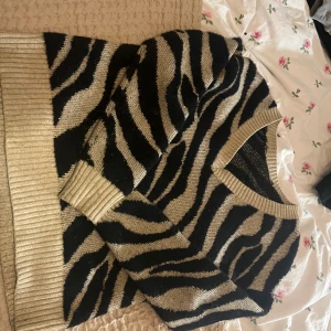 Zebramönstrad stickad tröja - Säljer en snygg zebramönstrad stickad tröja i svart och beige. Tröjan har en v-ringad hals och långa ärmar med ribbade muddar. Perfekt för att ge din outfit ett djärvt uttryck!