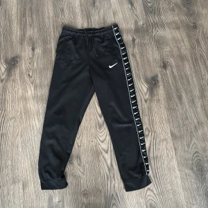 Svarta träningsbyxor från Nike - Snygga svarta träningsbyxor från Nike med elastisk midja och snörning. Byxorna har vita Nike-loggor längs sidorna och en liten logga framtill. Perfekta för träning eller en avslappnad dag. PRIS KAN DISKUTERAS!