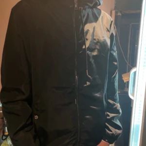 Svart vindjacka från Moncler storlek S - Snygg svart vindjacka från Moncler med huva och dragkedja. Jackan har en diskret logga på ärmen och är perfekt för blåsiga dagar. Den har en stilren design med en liten detalj i form av en röd och vit rand på insidan av huvan.