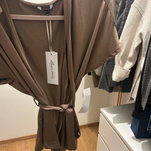 Brun omlottblus - Elegant brun omlottklänning från By Malene Birger med korta ärmar och knytband i midjan. Perfekt för en stilren look.