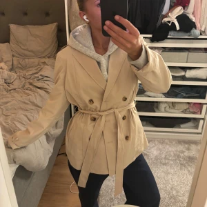 Beige trenchcoat med bälte - Snygg beige trenchcoat med knappar och bälte i midjan. Perfekt för höst och vår. Jackan har långa ärmar och en klassisk krage.