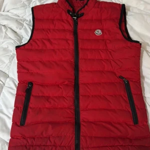 Röd dunväst moncler med dragkedja - Snygg röd dunväst i mycket bra skick. Perfekt för höst och vår, med praktiska dragkedjor på fickorna. Västen har en normal passform och är idealisk för lager-på-lager-stil. Den är quiltad för extra värme och komfort.