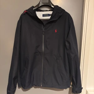 Svart vindjacka från Polo Ralph Lauren - Säljer en snygg svart vindjacka från Polo Ralph Lauren. Den har en dragkedja framtill och en liten röd logga på bröstet. Perfekt för höst och vår! Jackan har en huva och är i mycket bra skick. Passar både till vardags och lite finare tillfällen. Jackan är sprilands ny då den va lite för stor för mig. Köpt för 3000