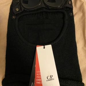 Svart stickad balaclava från C.P. Company - Säljer en cool svart stickad balaclava från C.P. Company med inbyggda glasögon. Perfekt för vintern och ger en unik look. Den är i nytt skick och har en tight passform. Glasögonen är fastsatta med knappar och kan justeras. Perfekt för att hålla sig varm och stilren under kalla dagar.