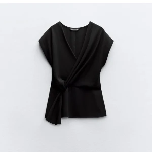 Svart omlottopp från Zara - Elegant svart omlottopp från Zara med en stilren design. Toppen/blusen har en v-ringad hals och knytning i sidan för en smickrande passform. Perfekt för en chic och modern look. Obs: finns ett mellanrum längst upp på dragkedjan så man kan stänga 90%! Inget som märks när man har på den eller som man tänker på. Skriv om du ha frågor