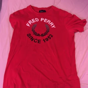 Röd t-shirt från Fred Perry - Snygg röd t-shirt från Fred Perry med tryck 'Since 1952' och den klassiska lagerkransen på bröstet. Perfekt för en casual look.