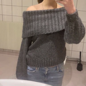 Grå offshoulder stickad tröja - Mysig grå stickad tröja med offshoulder-design. Perfekt för kyliga dagar med sin ribbade struktur och långa ärmar. En stilren och bekväm tröja som passar till jeans eller kjol.