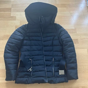 Svart pufferjacka från Esprit - Snygg svart pufferjacka från Esprit med dragkedja och huva. Jackan har ett bälte i midjan för en mer definierad passform och två praktiska fickor med dragkedja framtill. Perfekt för kyliga dagar.