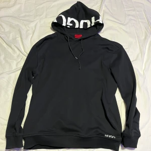 Svart hoodie från Hugo - Säljer en snygg svart hoodie från Hugo med vit text på huvan. Perfekt för både vardag och chill. Den har en klassisk passform med långa ärmar och en justerbar huva. Pris går att diskutera och byten går också 