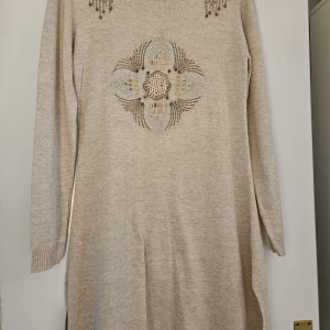 Beige stickad tunic med pärlor och paljetter - Säljer en superfin beige stickad tunic med ett detaljerat mönster av pärlor och paljetter på bröstet och axlarna. Perfekt för att glamma upp vardagen eller till festliga tillfällen. Tröjan har långa ärmar och en bekväm passform. Passar perfekt till höst och vinter!