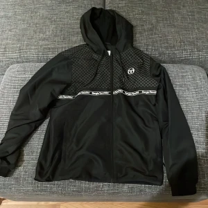 Svart jacka från Sergio Tacchini - Säljer en svart jacka från Sergio Tacchini i mycket bra skick. Den har en dragkedja framtill och ett snyggt rutmönster på axlarna. Jackan är långärmad och har en huva, perfekt för höst och vår. Den är gjord i ett syntetmaterial och har märkets logga tryckt på bröstet och längs med bröstet. Pris går alltid att förhandlas!