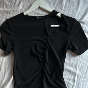 Svart topp från New yorker  - Snygg svart topp från New yorker i storlek M. Den har en unik design med en asymmetrisk detalj vid halsen och är tillverkad i ett mjukt material. Perfekt för både vardag och fest!