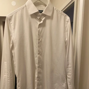 Vit skjorta från Dressmann - Säljer en klassisk vit skjorta från Dressmann i premium bomullsstretch. Skjortan har långa ärmar och knappar framtill. Perfekt för en stilren look.