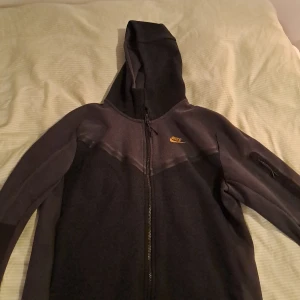 Svart och grå hoodie från Nike - Säljer en snygg svart och grå hoodie från Nike med dragkedja och huva. Perfekt för en sportig look eller avslappnade dagar. Den har en liten ficka på ärmen och det klassiska Nike-loggan på bröstet.(tech fleec) bara höra av sig vid funderingar 