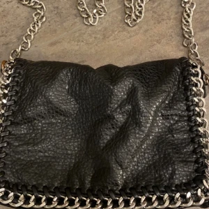 Svart axelväska med kedjedetaljer - Snygg svart axelväska från Tiamo i skinn med coola kedjedetaljer. Väskan har en dragkedja på baksidan och en kedja som axelrem. Perfekt för en kväll ute eller en dag på stan! NYTT RPIS 400