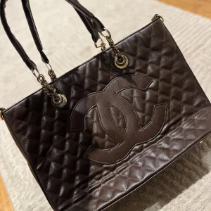 Brun quiltad handväska - Snygg brun quiltad handväska med kedjedetaljer och stora öglor. Väskan har ett stort logotypmärke på framsidan och är perfekt för att bära med sig alla nödvändigheter.