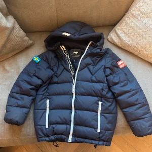 Mörkblå dunjacka från Mountain Works - Säljer en mörkblå dunjacka från Mountain Works med huva och dragkedja. Perfekt för kalla dagar! Passar för barn ca 7-9 år. I gott skick. Endast enstaka skråma. 