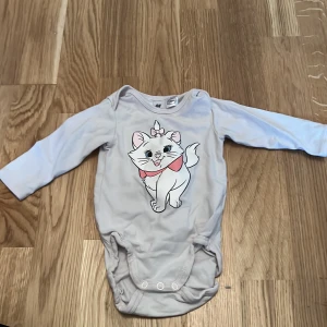 Vit långärmad body med kattmotiv från H&M - Söt vit långärmad body från H&M med ett gulligt kattmotiv från Disney. Tillverkad i ekologisk bomull, perfekt för de minsta. Praktiska knappar i grenen för enkel på- och avklädning.
