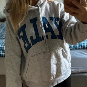 Grå hoodie med Yale-tryck - Säljer en superbekväm grå hoodie med stort blått Yale-tryck på framsidan. Perfekt för mysiga dagar eller casual outfits. Den har en klassisk känguruficka och justerbar huva med snören. Passar perfekt till jeans eller leggings!