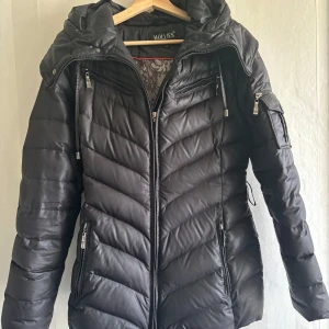 Svart dunjacka från Hollies - Säljer en snygg svart dunjacka från Hollies i storlek 34. Jackan har en dragkedja framtill och flera praktiska fickor med dragkedjor. Perfekt för kalla vinterdagar med sin varma och quiltade design. Huvan är avtagbar och ger extra skydd mot väder och vind. Passar både till vardags och lite finare tillfällen.