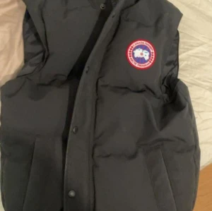 Svart dunväst från Canada Goose - Säljer en svart dunväst från Canada Goose i mycket bra skick. Västen har en klassisk design med knappar och det ikoniska märket på bröstet. Perfekt för kyliga höst- och vinterdagar. Håll dig varm och stilren! Köpt från NK för 7299kr priset kan diskuteras vid snabb affär