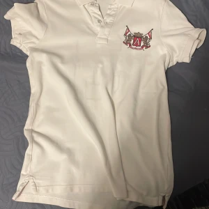 Vit pikétröja med emblem och nummer - Snygg vit pikétröja med ett broderat emblem på bröstet och en stor siffra '3' på ryggen. Tröjan har korta ärmar och en klassisk krage med knappar. Perfekt för en sportig look!