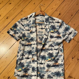 Mönstrad kortärmad skjorta från Hampton Republic - Säljer en supercool kortärmad skjorta från Hampton Republic i storlek S. Skjortan har ett tropiskt mönster med palmer och stränder i blått och orange på en vit bakgrund. Perfekt för sommarens alla äventyr! Regular fit för en bekväm passform. 🌴☀️