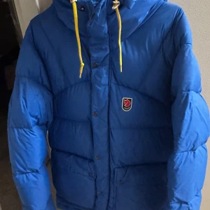 Blå dunjacka från Fjällräven - Säljer en blå dunjacka från Fjällräven, Expedition Series 1974. Jackan har gula snören vid huvan och knäppning med både dragkedja och knappar. Den är nytvättad därför den är i en påse. 