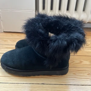 Svarta boots med päls från UGG - Säljer ett par svarta UGG boots i mycket bra skick. De är gjorda av mocka och har en mysig pälsdetalj runt skaftet. Perfekta för kalla vinterdagar när du vill hålla fötterna varma och stiliga. Passar både till vardags och lite finare tillfällen.