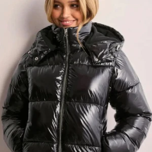 Svart glansig pufferjacka Nelly - Snygg svart pufferjacka med glansig yta och dragkedja framtill. Jackan har en huva och långa ärmar. Stl S