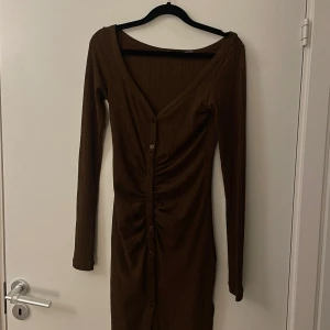 Brun ribbad klänning - Snygg brun ribbad klänning med v-ringning och knappar framtill. Den har långa ärmar och en figurnära passform som framhäver silhuetten. Perfekt för en stilren look.