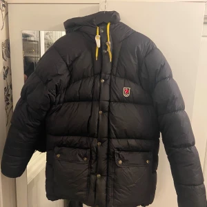 Svart dunjacka från Fjällräven - Säljer en svart dunjacka från Fjällräven med gula detaljer. Jackan har en huva och knappar framtill samt en broderad logotyp på bröstet. Perfekt för kalla dagar.