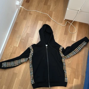 Svart kofta/tröja från Burberry - Säljer en skön och äkta svart huvtröja från Burberry med klassiskt rutigt mönster på ärmarna. Tröjan har en dragkedja framtill och en justerbar huva. Perfekt för en trendig och bekväm look