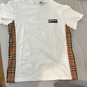 Vit t-shirt med Burberry-rutor - Snygg och äkta vit t-shirt från Burberry med klassiska rutiga paneler på sidorna. T-shirten har en Burberry-logga på bröstet och är tillverkad i mjukt bomullsmaterial. Perfekt för en sommar dag eller allmänt. T-shirten har aldrig använts förr.