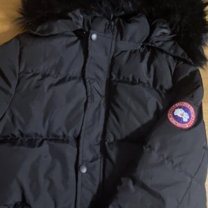 Svart dunjacka från Canada Goose - Säljer en svart dunjacka från Canada Goose i storlek S. Jackan har en pälskantad huva och är perfekt för kalla vinterdagar. Den har en dragkedja och knappar framtill samt det klassiska Canada Goose-märket på ärmen. Supervarm och stilren!