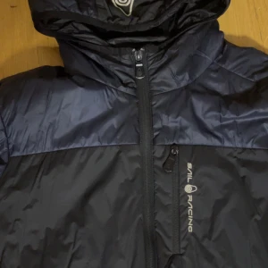 Svart och mörkblå jacka från Sail Racing - Säljer en svart jacka från Sail Racing i bra skick med ett litet hål på vänstra armen. Jackan har en dragkedja framtill och en justerbar huva. Perfekt för höst- och vårsäsongen. Den är tillverkad i ett syntetmaterial som ger bra skydd mot vind och kyla. Passar både till vardags och utomhusaktiviteter.