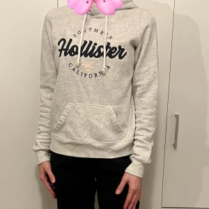 Grå hoodie från Hollister - Säljer en supermysig grå hoodie från Hollister med tryck 'Southern California' på bröstet. Perfekt för chilliga dagar eller som ett extra lager under jackan. Den har en praktisk magficka och justerbar huva med snören. Passar både till vardags och avslappnade tillfällen.