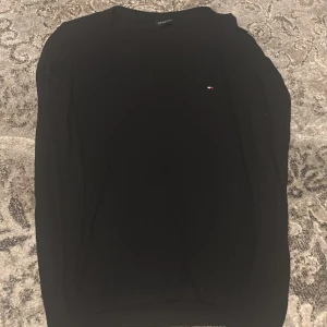 Svart tröja från Tommy Hilfiger - Snygg svart tröja från Tommy Hilfiger med diskret logga på bröstet. Tröjan har en v-ringad hals och är perfekt för en stilren look.