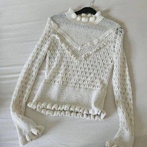 Vit stickad tröja med volangdetaljer - Säljer en elegant vit stickad tröja med vackra volangdetaljer vid hals, ärmslut och nederkant. Tröjan har ett fint mönster och långa ärmar, perfekt för en stilren look. Passar utmärkt till både vardag och speciella tillfällen.