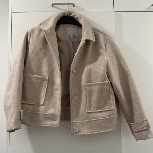 Beige kappa från Arket - Snygg beige kappa från Arket med stora fickor och knappar framtill. Jackan har en klassisk krage och är perfekt för höst och vår. Den har en stilren och tidlös design som passar till många olika outfits.