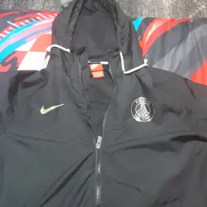 Säljer en svart Nike vindjacka i storlek M med PSG-logga på bröstet. Jackan har en dragkedja framtill och en justerbar huva med vita snören. Perfekt för höst och vår när du vill hålla stilen och samtidigt visa ditt stöd för Paris Saint-Germain. Köpte den från nike sidan helt ny den är använd bara 2 gånger