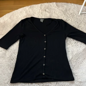 Svart ribbad kofta från Lindex - Säljer en stilren svart ribbad kofta från Lindex i storlek S. Den har trekvartsärmar och knappar framtill, perfekt för både vardag och fest. Koftan är i mycket bra skick och passar till alla säsonger. Ett hållbart val för din garderob!