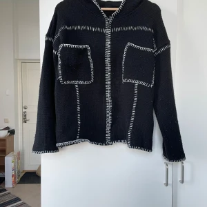 Svart stickad kofta från BDG - Säljer en svart stickad kofta från BDG med vita sömmar som ger en cool kontrast. Koftan har långa ärmar och en avslappnad passform. Perfekt för höst och vinter när du vill ha något bekvämt och stilrent. Den har en dragkedja framtill och två praktiska fickor.