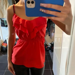 Volangtopp - Säljer en snygg röd ärmlös topp med volangdetaljer framtill. Toppen har en smickrande passform med resår i midjan och tunna axelband. Perfekt för sommarens fester eller en utekväll. Den är i mycket bra skick och redo för nya äventyr! 💃