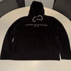 Säljer en klassisk svart hoodie från Tommy Hilfiger med logotypen tryckt på bröstet. Den har en bekväm passform och en praktisk magficka. Perfekt för en avslappnad stil.