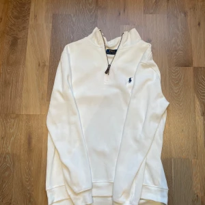 Vit Quarter Zip från Ralph Lauren - Säljer en stilren vit tröja från Polo Ralph Lauren med dragkedja och det klassiska logotypbroderiet på bröstet. Modellen är 170cm och väger 65kg. Skriv om ni har frågor!