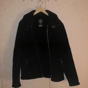 Svart jacka från King x JoeMia - Säljer en svart jacka från King x JoeMia i storlek 48.Jackan har en dragkedja framtill och en snygg bältesdetalj vid kragen. Perfekt för höst och vinter med sitt varma material.