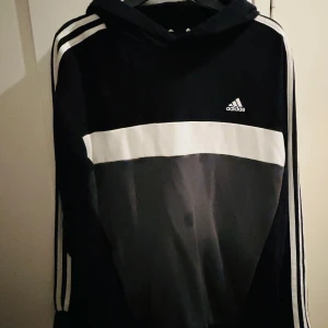 Svart och vit hoodie från Adidas storlek 176 - Säljer en snygg svart och vit hoodie från Adidas i storlek 176 som motsvarar nästa storlek S. Den har en klassisk design med tre ränder längs ärmarna och en stor vit rand över bröstet. Perfekt för både träning och vardag. Huvan ger extra mysfaktor. 